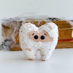 May include: A plush tooth dressed as a mummy for Halloween. The tooth is wrapped in white fabric bandages, with black button eyes and a light pink base. Spiders and cobwebs are in the background.