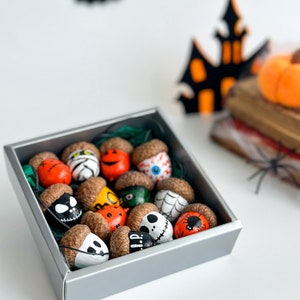 K&ouml;nnte beinhalten: Eine graue Box gef&uuml;llt mit Eicheln, die mit verschiedenen Halloween-Motiven bemalt sind, darunter ein Geist, eine Spinne, ein K&uuml;rbis und Jack Skellington. Die Eicheln befinden sich in einer Box auf einer wei&szlig;en Oberfl&auml;che.