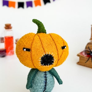 May include: A felt pumpkin character with a green stem, orange body, and a black stitched mouth. The character is wearing a blue felt body suit with stitched seams.