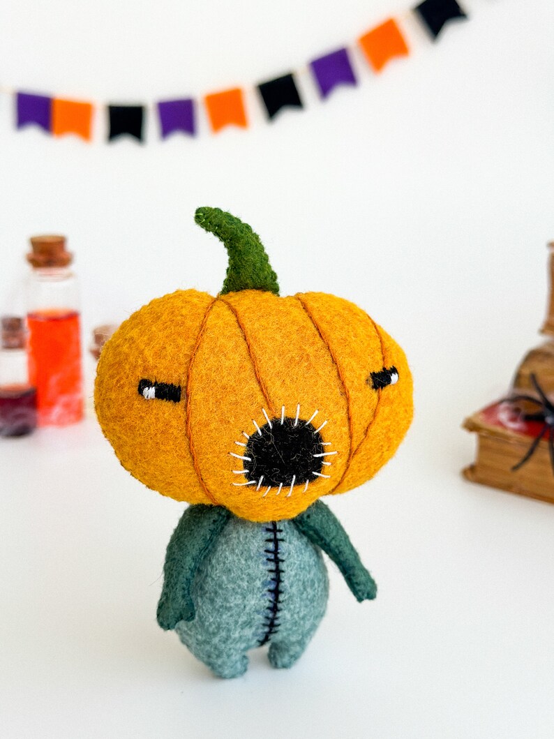 May include: A felt pumpkin character with a green stem, orange body, and a black stitched mouth. The character is wearing a blue felt body suit with stitched seams.