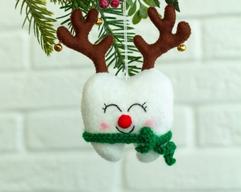 May include: A white felt tooth ornament with a red nose and green scarf, decorated with brown felt antlers and gold bells. The ornament is hanging from a green branch with red berries.