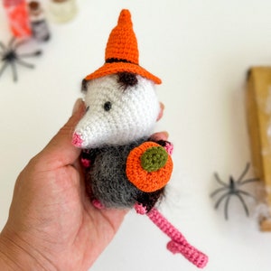 May include: A crocheted opossum wearing a witch hat and holding a small orange pumpkin. The opossum is white with black eyes and a gray body. The hat is orange with a black brim. The pumpkin is orange with a green stem.