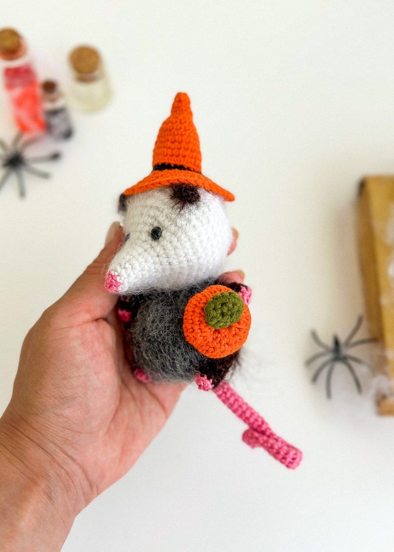 May include: A crocheted opossum wearing a witch hat and holding a small orange pumpkin. The opossum is white with black eyes and a gray body. The hat is orange with a black brim. The pumpkin is orange with a green stem.