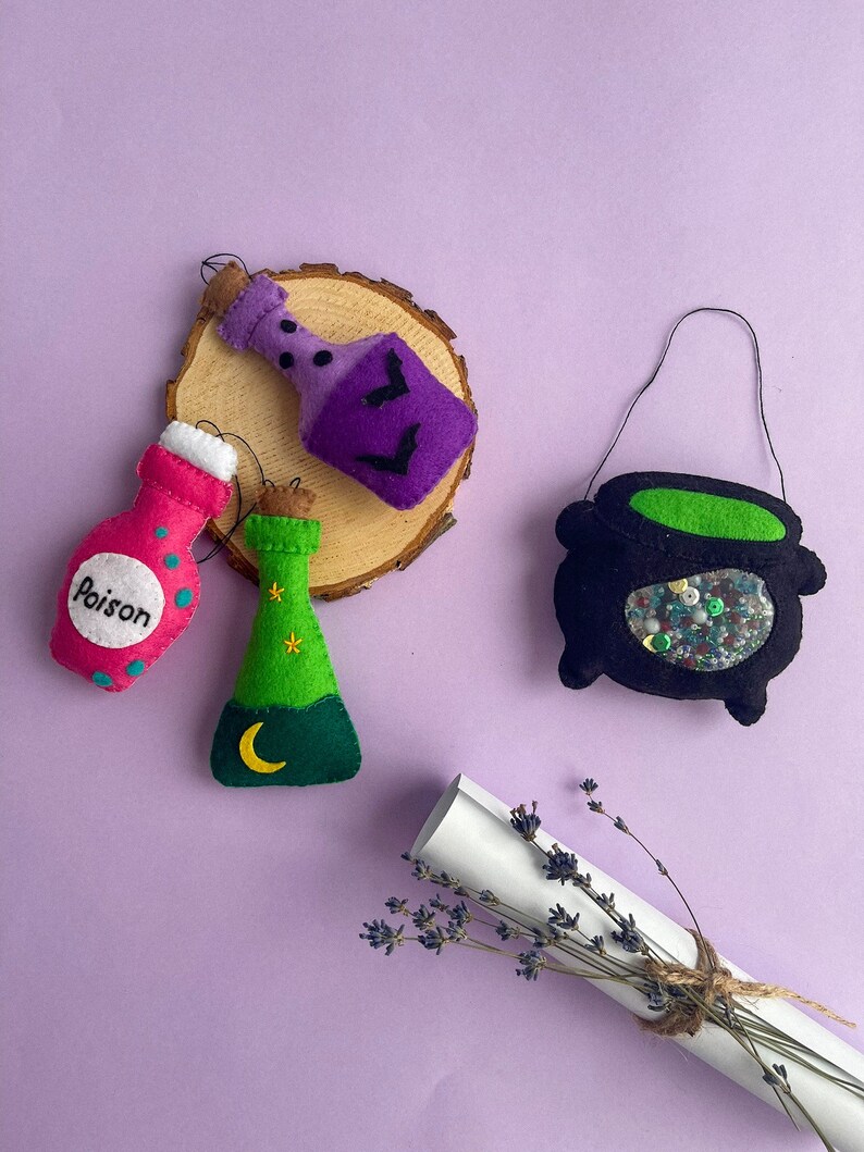 Puede incluir: Un conjunto de decoraciones de Halloween de fieltro que incluye un caldero, una botella de poci&oacute;n morada, una botella de poci&oacute;n verde con una luna creciente y estrellas, y una botella rosa que dice "Poison".