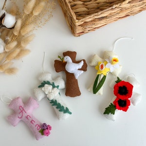 Puede incluir: Una colección de cruces de fieltro hechas a mano en varios colores, cada una adornada con adornos únicos. Una cruz es marrón con una paloma blanca, otra rosa con el nombre "Anna", y otras presentan flores.