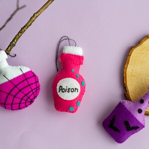 Puede incluir: Tres botellas de poci&oacute;n de Halloween de fieltro con diferentes dise&ntilde;os. Una botella es rosa con lunares blancos y dice "Poison". Otra botella es rosa con un dise&ntilde;o de telara&ntilde;a negra. La tercera botella es morada con un dise&ntilde;o de murci&eacute;lago.