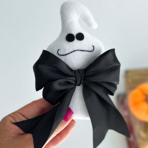 May include: White felt ghost with black eyes and a stitched-on smile, wearing a black satin bow tie.