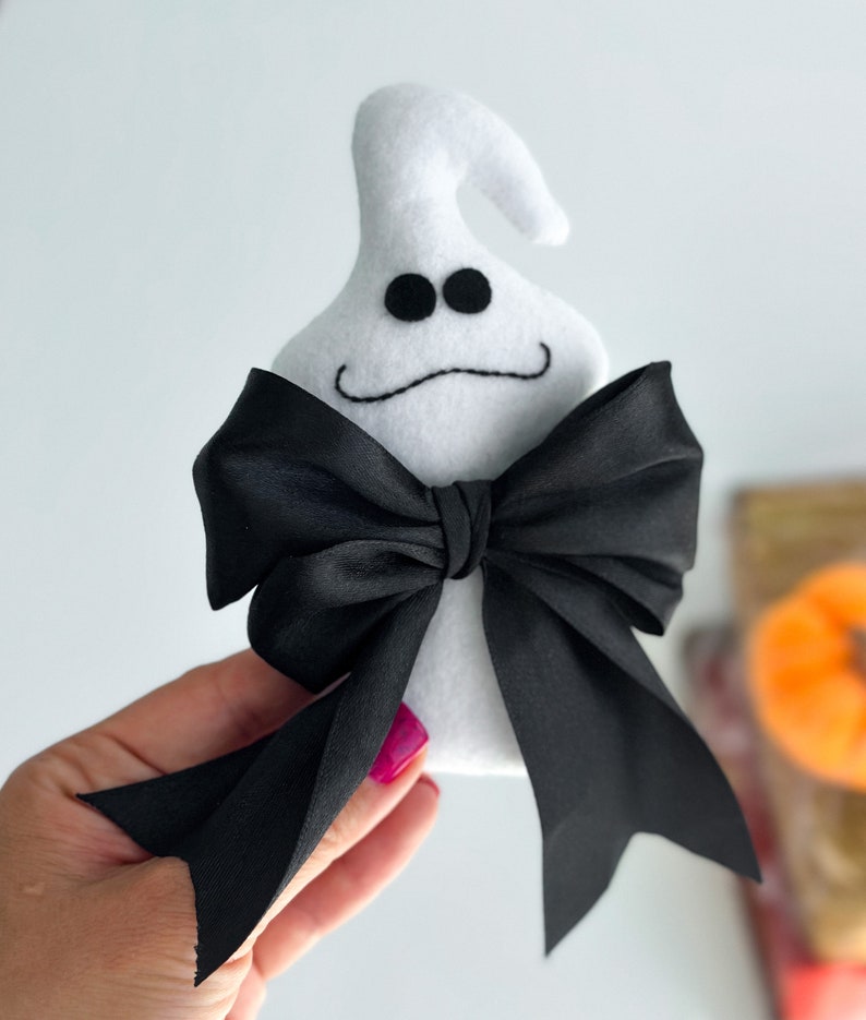May include: White felt ghost with black eyes and a stitched-on smile, wearing a black satin bow tie.
