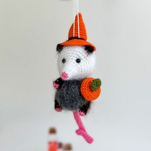 May include: A crocheted opossum wearing an orange witch hat and holding a small orange pumpkin. The opossum is hanging from a string.