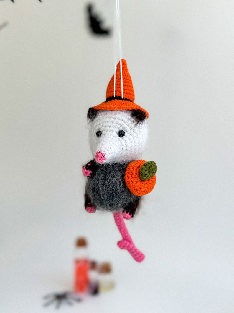 May include: A crocheted opossum wearing an orange witch hat and holding a small orange pumpkin. The opossum is hanging from a string.