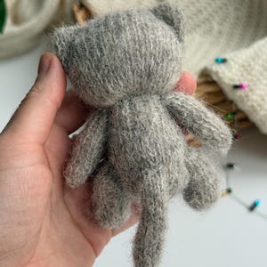 K&ouml;nnte beinhalten: Ein kleines, graues, gestricktes Katzenspielzeug mit Schwanz, Ohren und vier Beinen. Die handgefertigte Katze wird in der Hand einer Person gehalten. Die Katze ist aus einem weichen, flauschigen Material gefertigt.