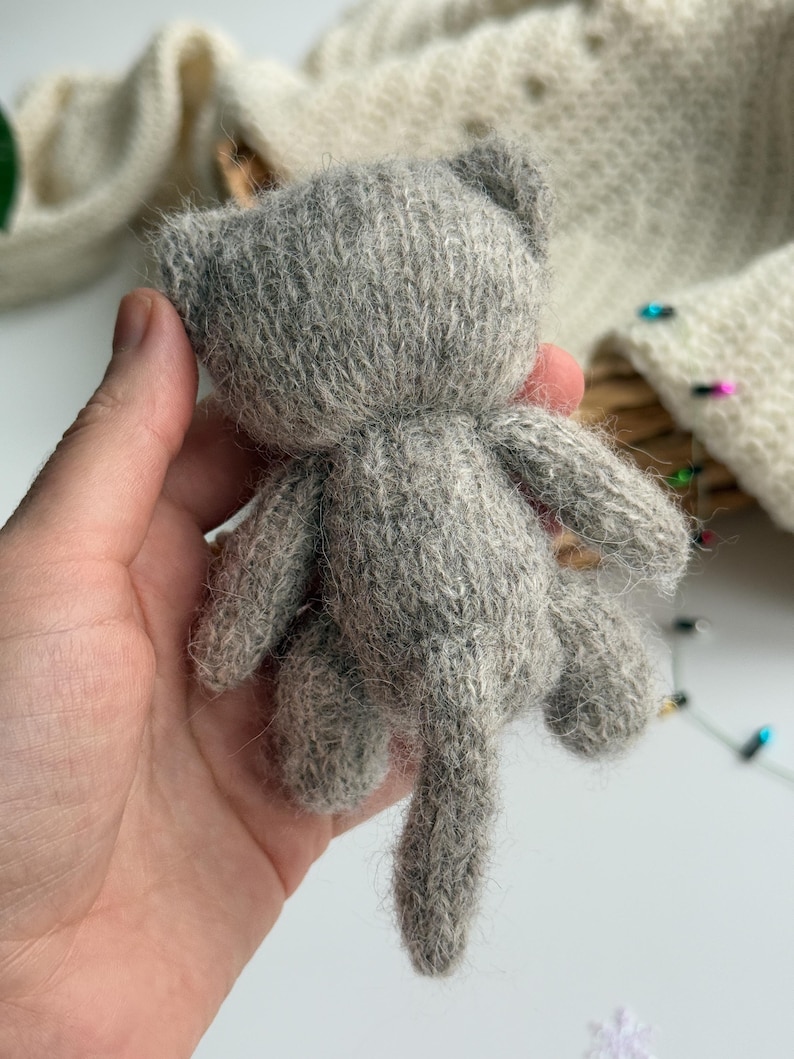 K&ouml;nnte beinhalten: Ein kleines, graues, gestricktes Katzenspielzeug mit Schwanz, Ohren und vier Beinen. Die handgefertigte Katze wird in der Hand einer Person gehalten. Die Katze ist aus einem weichen, flauschigen Material gefertigt.