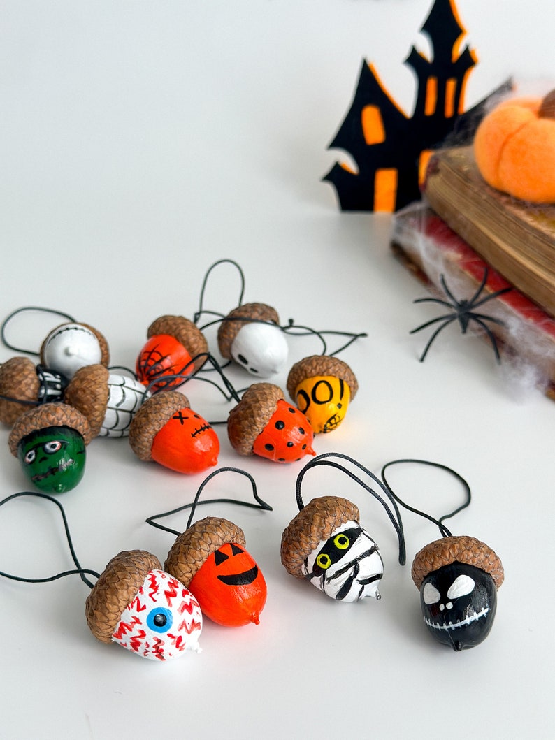 K&ouml;nnte beinhalten: Eine Sammlung von Eicheln, die mit Halloween-Motiven bemalt sind, darunter Geister, K&uuml;rbisse, Spinnen und Mumien. Die Eicheln sind mit schwarzem Garn aufgereiht und bereit, als Dekoration verwendet zu werden.