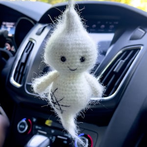 May include: A white crocheted ghost with a smiling face and a black spiderweb design hanging from the rearview mirror of a car.
