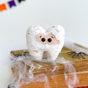 May include: A plush tooth wrapped in white bandages, resembling a mummy, with black button eyes. It rests on a stack of old books, decorated with spiderwebs and toy spiders, with a Halloween banner in the background.
