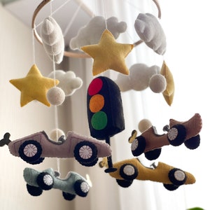 May include: A felt baby mobile with a traffic light, stars, clouds, and cars. The cars are in shades of brown, gray, and blue. The stars are yellow, and the clouds are white.