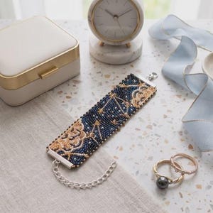 May include: A wide beaded bracelet featuring a dark blue background with gold and white celestial patterns, including star constellations. The bracelet has silver-toned metal end bars and a silver chain extension with a lobster claw clasp closure.