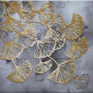 Könnte beinhalten: Wandkunst mit einem Muster aus stilisierten Ginkgoblättern. Die Blätter sind aus goldenen und cremefarbenen Perlen gefertigt, wodurch ein strukturierter, dreidimensionaler Effekt entsteht, vor einem grauen Hintergrund mit Blattformen.