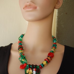 May include: A green, red, and gold glass bead necklace with a large green, red, and gold glass pendant. The necklace is on a mannequin wearing a black shirt.