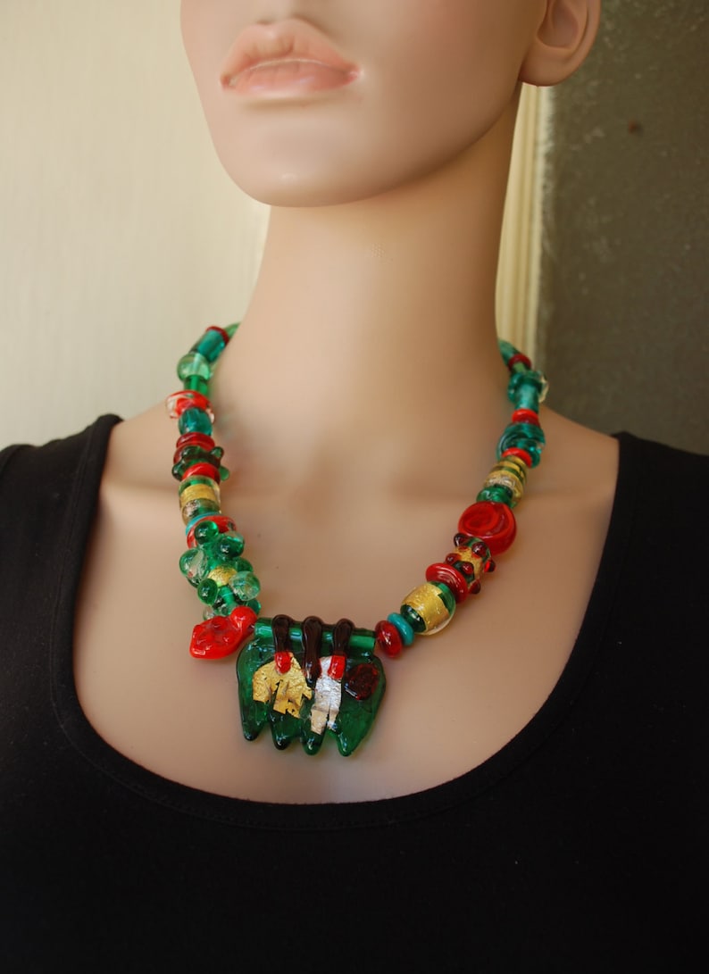 May include: A green, red, and gold glass bead necklace with a large green, red, and gold glass pendant. The necklace is on a mannequin wearing a black shirt.