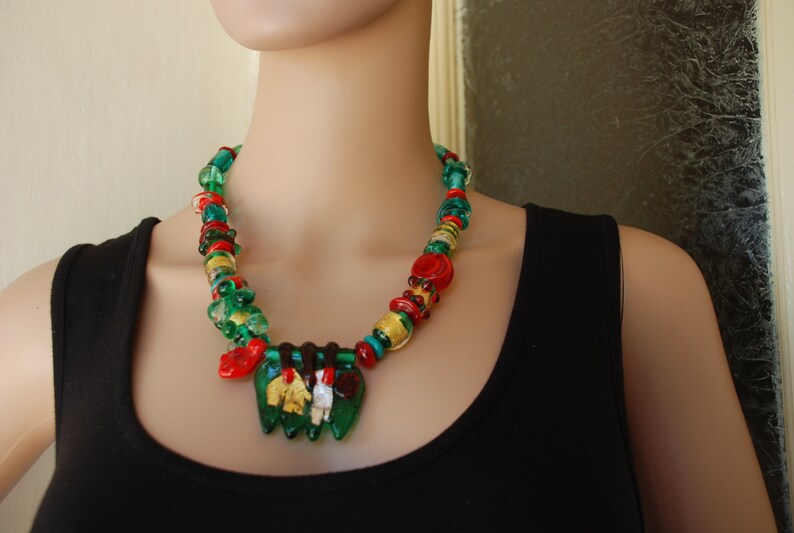 May include: A multi-colored glass bead necklace with green, red, and gold beads. The necklace has a pendant with a green, gold, and red design.