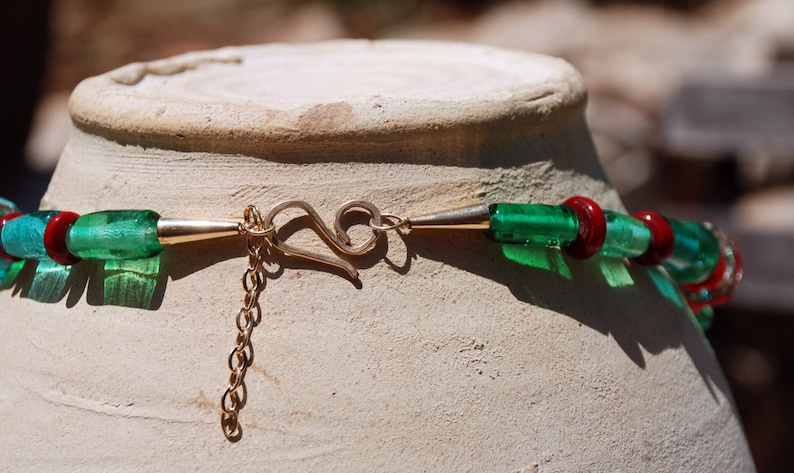 May include: A close-up of a green and red glass bead necklace with a gold clasp. The necklace is lying on a white ceramic surface.