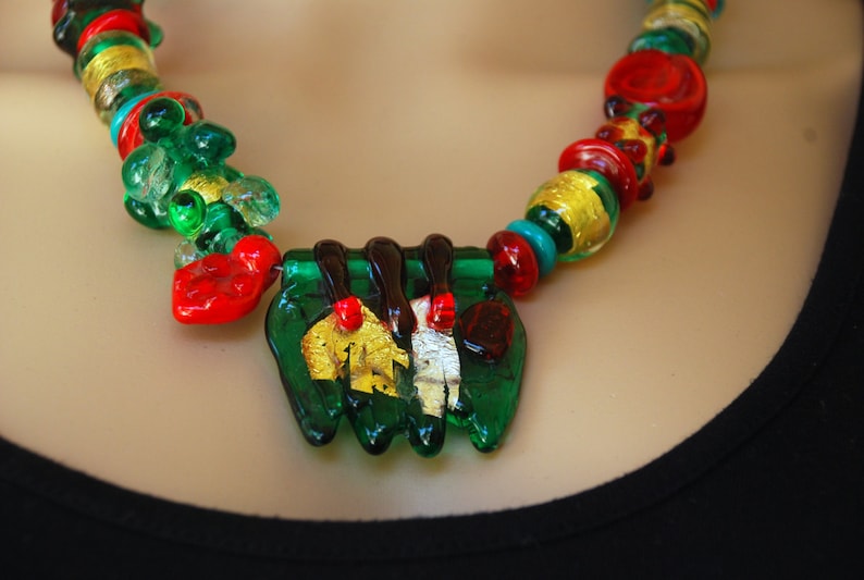 May include: A green, red, and gold glass necklace with a large green pendant featuring gold leaf accents.