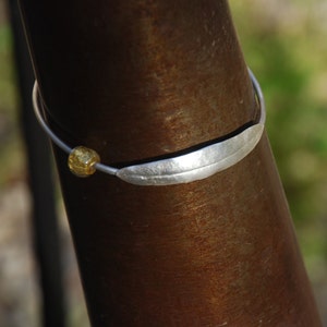 May include: A silver bracelet with a leaf-shaped charm and a gold bead. The bracelet is on a brown metal surface.