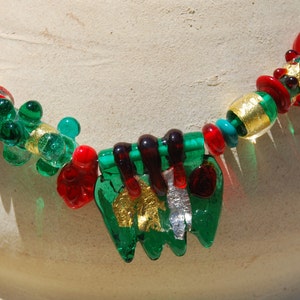 May include: A green, red, and gold glass necklace with a unique, multi-layered pendant. The pendant features a green, leaf-like design with gold accents.