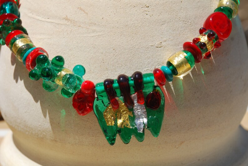 May include: A green, red, and gold glass necklace with a unique, multi-layered pendant. The pendant features a green, leaf-like design with gold accents.