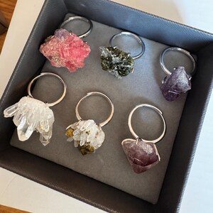 May include: A collection of six rings with raw crystal clusters in a grey box. The rings feature various stones including a pink mineral, a dark green textured cluster, a deep purple crystal, a clear quartz cluster, a white and gold mineral, and a dark purple stone.