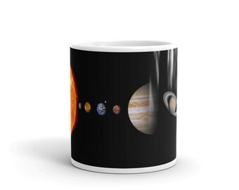 May include: A white ceramic mug with a black exterior featuring a graphic print of the solar system. The design displays the sun, Mercury, Venus, Earth, Mars, Jupiter, and Saturn arranged in a horizontal line against a black background.