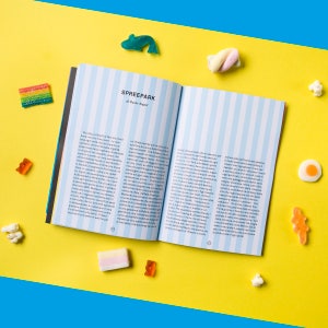 May include: An open book with blue and white striped pages. The book is titled "Spreepark" and the text on the pages is in Italian. The book is lying open on a yellow surface with colourful candy scattered around it.