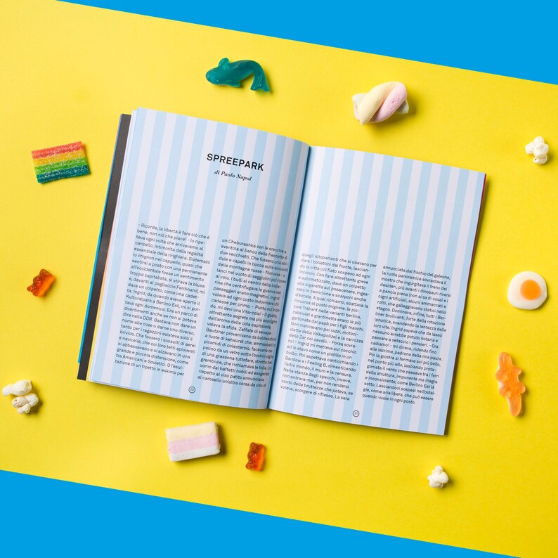 May include: An open book with blue and white striped pages. The book is titled "Spreepark" and the text on the pages is in Italian. The book is lying open on a yellow surface with colourful candy scattered around it.