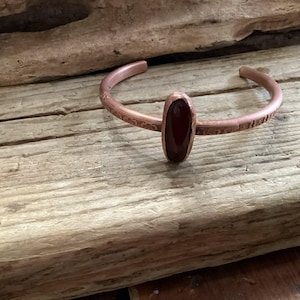 May include: A copper cuff bracelet with an oval, reddish-brown stone centerpiece. The bracelet has a textured design and is displayed on a weathered wooden surface. The jewelry is a handcrafted accessory.
