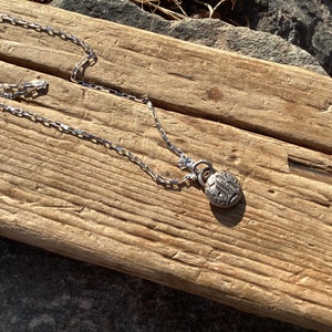 May include: A silver chain necklace with a round pendant featuring an intricate design. The necklace is displayed on a piece of weathered wood.
