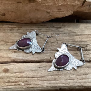 May include: A pair of dangle earrings featuring faceted, oval-shaped deep red stones set in textured, hammered silver-toned metal. The metal components have an irregular, organic triangular shape with a dark, oxidized finish and thin wire hooks.