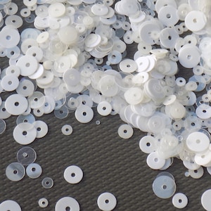 May include: A large pile of white, round sequins. The sequins are scattered on a dark grey surface.