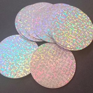 May include: A pile of iridescent white round sequins with a hole in the centre. The sequins are made of a shiny material that reflects light.