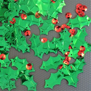 以下が含まれることがあります： 緑色のヒイラギの葉の形をしたスパンコールと、赤いスパンコールのベリーが灰色の表面に散らばっているクローズアップ。これらの光沢のあるお祝いの装飾は、ホリデーの工芸品、スクラップブッキング、またはあらゆるプロジェクトに輝きを加えるのに最適です。