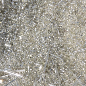 May include: A close-up of a pile of small, shiny, silver seed beads. The beads are a mix of round and tube shapes, creating a textured surface. The light reflects off the beads, giving them a bright, metallic appearance. Suitable for jewellery making.