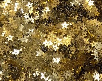 Op de afbeelding: Een close-up afbeelding van een grote hoeveelheid gouden stervormige pailletten. De pailletten zijn willekeurig verspreid en creëren een glinsterend, sprankelend effect.