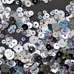 May include: A close-up of a variety of iridescent sequins in silver, black, and blue. The sequins are scattered on a black surface.