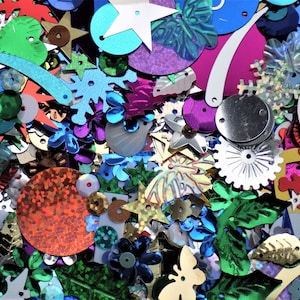 May include: A colourful collection of sequins in diverse shapes such as stars, flowers, circles, and leaves. The sequins are in shades of blue, green, gold, silver, purple, and red, creating a dazzling, textured surface.