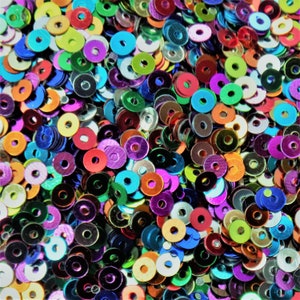 May include: A large assortment of colourful iridescent sequins in various shades of blue, green, purple, red, orange, yellow, and silver. The sequins are all round and have a metallic sheen.