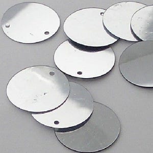 May include: A selection of silver, circular sequins. Each disc has a small hole near the edge. The sequins are scattered on a light grey surface, reflecting light and creating a bright, metallic appearance. Suitable for craft projects and embellishments.