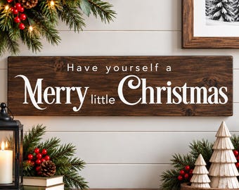 May include: A rectangular dark brown wooden sign with white text that reads Have yourself a Merry little Christmas. The sign is displayed with evergreen branches, red berries, a black lantern with a candle, and small wooden tree figures.