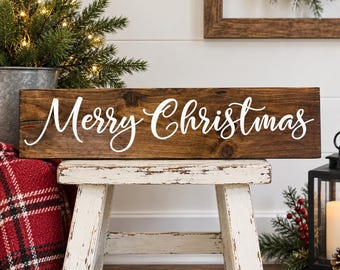 May include: A rectangular dark brown wooden sign featuring the white script text Merry Christmas. The sign is positioned horizontally on a white wooden stool, showcasing a natural wood grain texture and visible knots across the surface.
