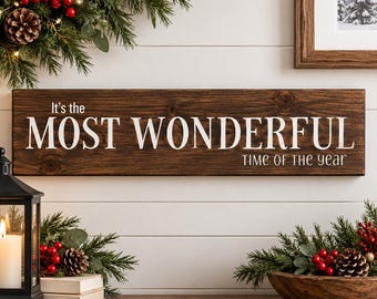May include: A rectangular dark brown wooden sign with white lettering that reads It's the MOST WONDERFUL TIME OF THE YEAR. The sign is displayed horizontally against a white paneled wall, positioned between holiday greenery and a lantern.