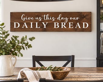 May include: A rectangular dark brown wooden wall sign featuring the white text Give us this day our DAILY BREAD. The sign has a smooth finish and is mounted horizontally on a plain wall above a dining table setting with a white pitcher and bowl.